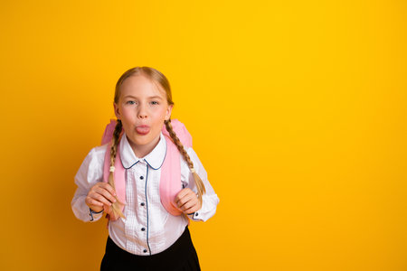 Adorable blonde schoolgirl with braids posing playfully with a pink backpack against a vibrant yellow backgroundの写真素材