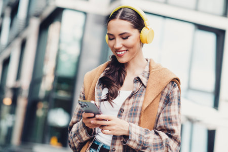 Young woman outdoors in an urban setting enjoying music with yellow headphones and a smartphone,...