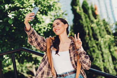 Cheerful young woman taking a selfie outdoors in a trendy casual outfit on a sunny day, enjoying...