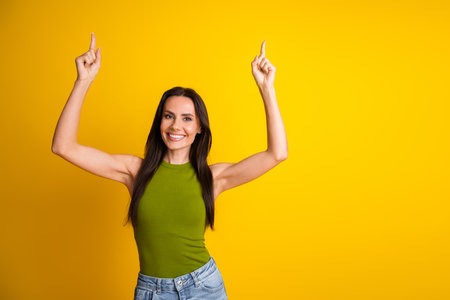 Smiling woman pointing upward on yellow background, wearing casual khaki top and jeans, expressing happiness and confidenceの写真素材