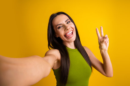 Excited young woman takes a cheerful selfie showing peace sign against a yellow background wearing a green topの写真素材