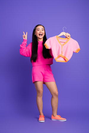 Smiling young woman holding colorful fashion clothing on a hanger, dressed in pink, standing against a purple vertical backgroundの写真素材