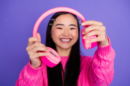 Cheerful young woman holding vibrant pink headphones, showcasing trendy fashion style in casual attire against purple backdropの写真素材