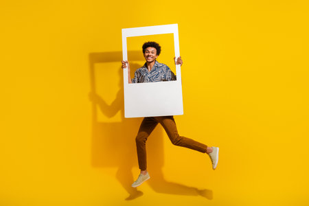 Portrait of a young man holding a large photo frame while joyfully jumping against a vibrant yellow backgroundの写真素材