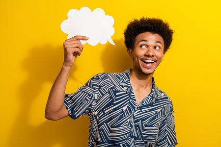 Happy young man holding a speech bubble on a bright yellow background, smiling cheerfully in casual...