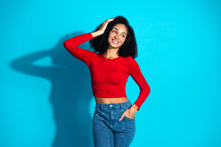 Confident young woman in a red top and jeans posing against a bright blue background, embracing a stylish casual vibeの写真素材