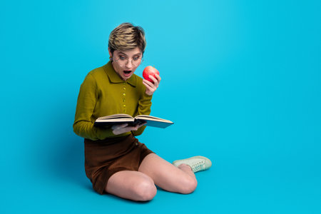 Young woman with short hair reads a book while holding an apple against a bright blue background