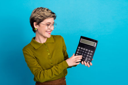 Smiling young woman with glasses holding a large calculator on a blue background in cheerful casual styleの写真素材