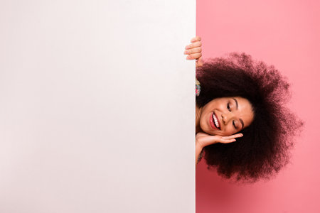 Positive woman with afro hairstyle peeking behind a blank white panel on a pink background, smiling cheerfully in stylish attireの写真素材