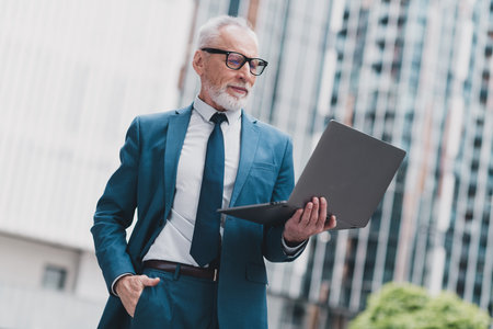 Confident senior businessman working on a laptop outdoors in an urban setting, standing near modern buildings.の写真素材