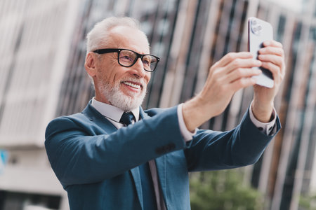 Successful mature businessman smiling while taking a selfie outdoors in a modern urban setting on a sunny dayの写真素材