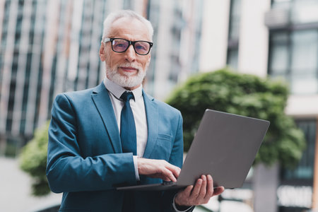 Confident mature businessman with laptop working outdoors showcasing professionalism, entrepreneurship, and urban office lifestyleの写真素材