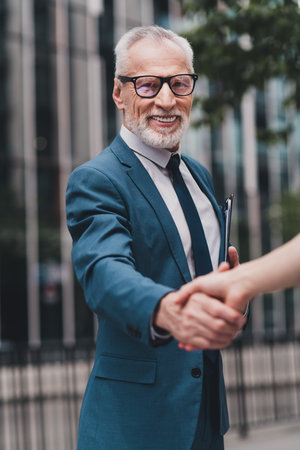 Successful senior businessman in formal suit shaking hands outdoors in a city environment, representing professionalismの写真素材