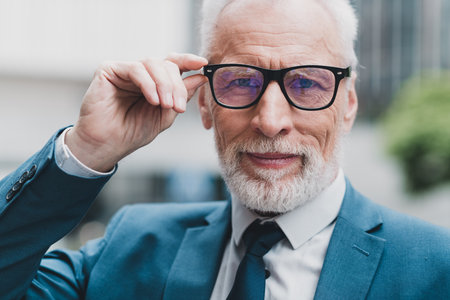 Confident senior businessman adjusting glasses outdoors, embodying success in urban settings, dressed in stylish professional attireの写真素材