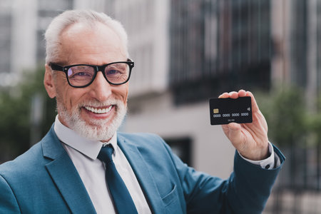 Confident senior businessman holding a black credit card outdoors in an urban environment while smiling professionallyの写真素材