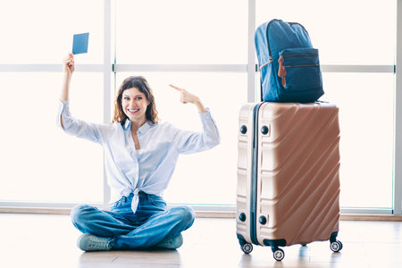 Young woman sitting with luggage and pointing to her boarding pass at the airport terminal, ready for travelの写真素材