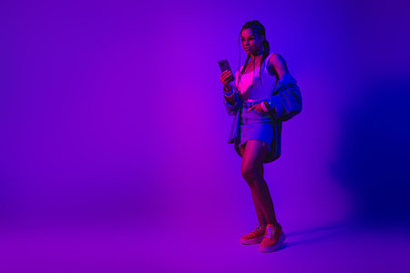Stylish young woman holding a phone in neon lighting, wearing denim and colorful outfit, exuding...