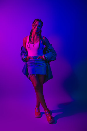 Young woman posing in neon lights wearing denim skirt and trendy jacket showcasing modern fashion...