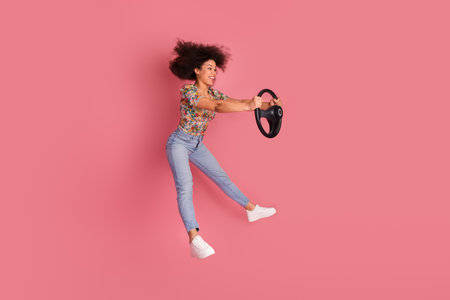 Young woman holding a steering wheel against a vibrant pink background modeling a stylish casual outfit and carefree energyの写真素材