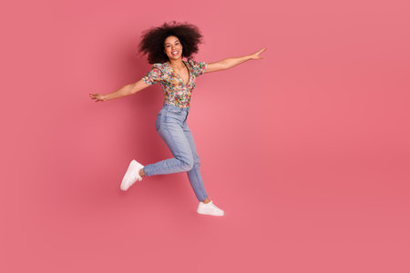 Happy young woman jumping joyfully on a pink background wearing casual stylish jeans and a floral...