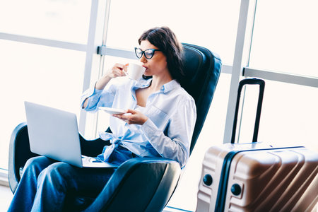 Young woman in casual fashion working on laptop while enjoying coffee at airport lounge during travelの写真素材