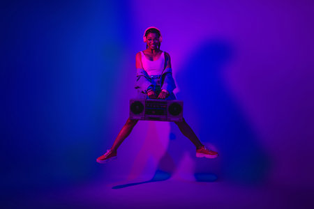 Stylish young woman holding a boombox in a vibrant violet and neon-lit room, expressing fashionable...