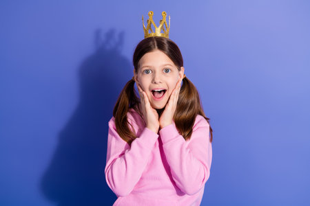Surprised young girl with a golden crown on purple background showing excitement with hands on her face in casual attireの写真素材