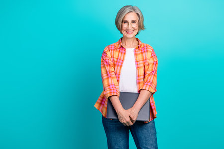 Smiling mature woman in casual outfit holding a laptop against a turquoise background, showcasing...
