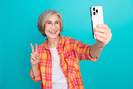 Happy mature woman taking selfie showcasing peace sign and charm in casual style against turquoise...