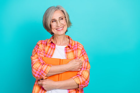 Charming elderly woman with grey hair wearing a stylish checkered shirt, holding an orange notebook, smiling confidentlyの写真素材