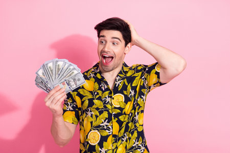 Cheerful young man holding dollar bills symbolizing wealth and success against a vibrant pink background in a tropical print shirtの写真素材