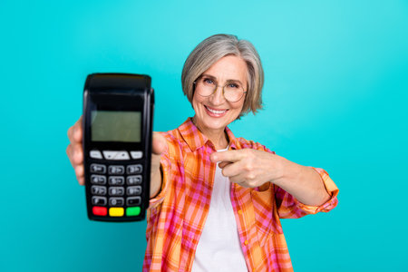 Senior woman holding payment terminal smiling confidently against teal background promoting cashless shopping and modern lifestyleの写真素材