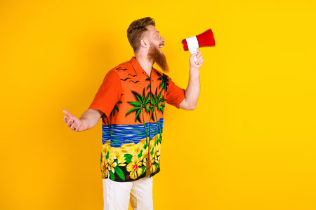 Vibrant young man with red beard wearing tropical shirt poses enthusiastically with megaphone, yellow backgroundの写真素材
