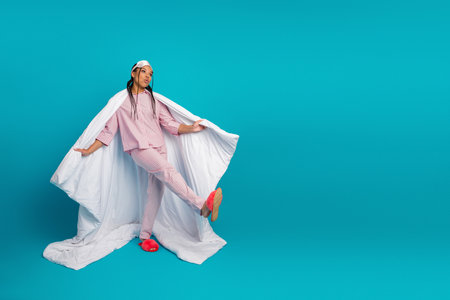 Young woman in pink pajamas with white duvet striking a playful pose against a cheerful blue background.の写真素材