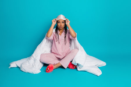 Young woman in pink pajamas with duvet and sleep mask sitting on blue background in a relaxed and playful pose.の写真素材
