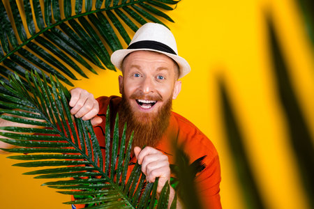 Happy red-haired man in hat smiling in tropical-themed setting on vibrant yellow background portraying summer vibesの写真素材