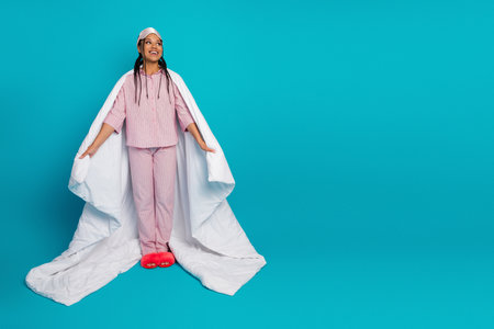 Charming young woman in pink sleepwear with duvet showcasing delightful comfort on vivid blue backgroundの写真素材