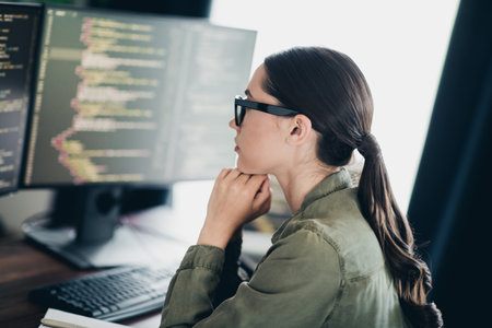 Charming female programmer working attentively in a home office environment on coding tasks with a computer monitorの写真素材