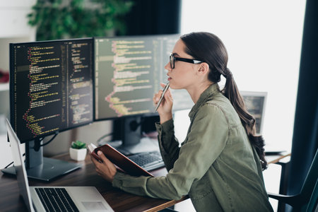 Focused young female programmer analyzing code on computers while working on laptop in an organized office spaceの写真素材