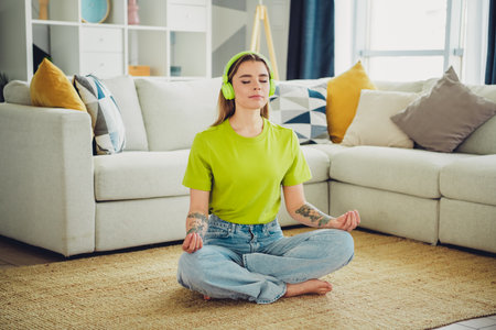 Young woman in casual attire meditates on a carpet in a cozy living room, enjoying a moment of relaxation and mindfulnessの写真素材
