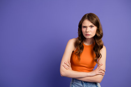 Charming young woman in casual orange top with arms crossed against purple backdrop, expressing curiosity and determinationの写真素材