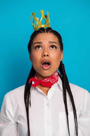 Adorable young woman in stylish casual wear with crown accessory expressing surprise against a blue backgroundの写真素材