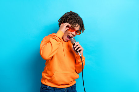 Energy-filled boy in orange hoodie singing and performing with microphone against vibrant blue backgroundの写真素材