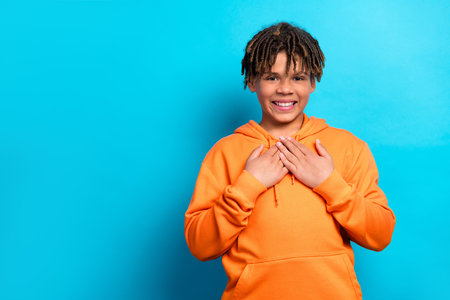 Cheerful schoolboy expressing gratitude in an orange hoodie against a vibrant blue background, displaying joyの写真素材