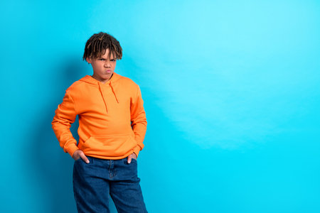 Young boy in stylish orange hoodie posing with curious expression against vibrant blue background representing casual attitudeの写真素材