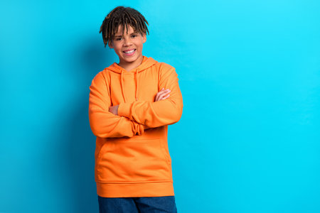 Cheerful teenage boy in vibrant orange hoodie crossing arms, standing confidently against bright blue backgroundの写真素材