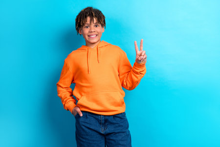 Cheerful boy in orange hoodie making peace gesture against blue backgroundの写真素材