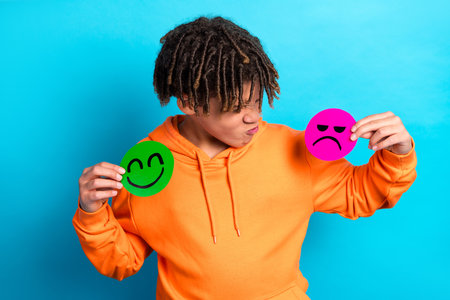 Young boy holding happy and sad emoji faces, expressing emotions and feelings against a vibrant blue...