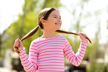 Smiling young girl in a pink striped shirt enjoying a bright sunny day outdoors, posing playfully while holding her hairの写真素材