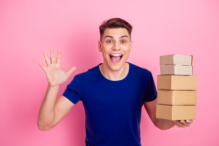 Cheerful young man holding cardboard boxes and waving with a joyful smile on a pink background
